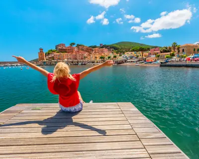 Tourist woman sitting over jetty and looking at Rio Marina harbor of Elba island in red suit. Lifestyle tourist girl on holiday travel in Italy, Europe. Rio Marina skyline cityscape on background,